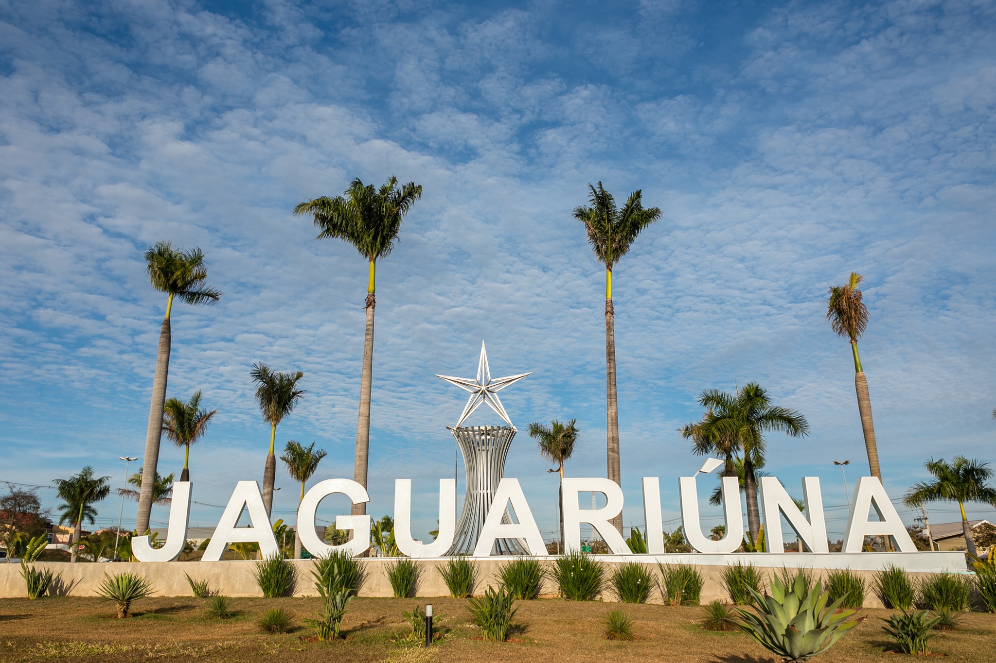 Área externa com letreiro 'JAGUARIUNA' em grandes letras brancas, escultura prateada em forma de estrela ao centro, palmeiras e céu azul com nuvens.
